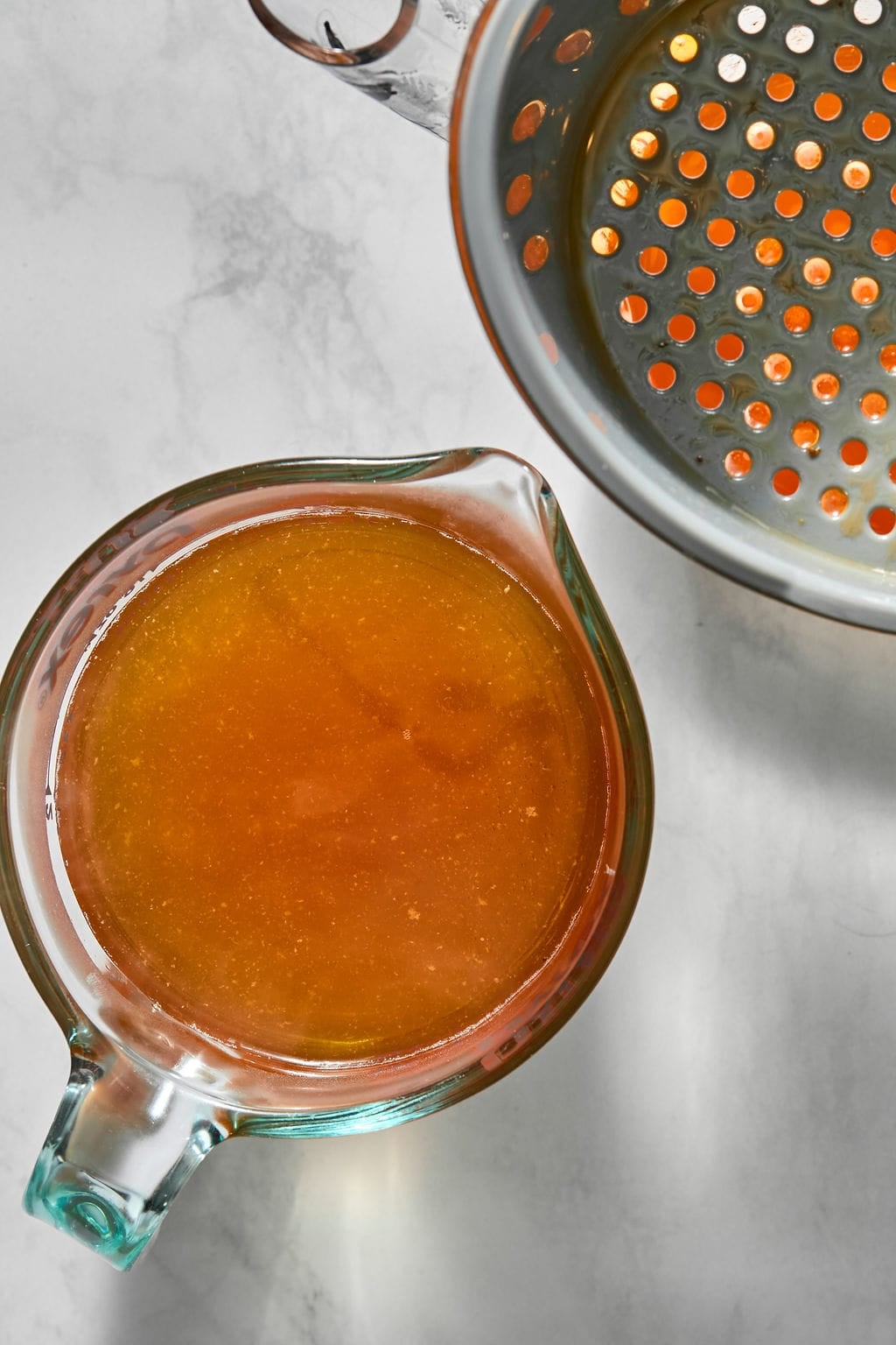 A glass measuring cup filled with golden-brown liquid next to a stainless steel strainer.