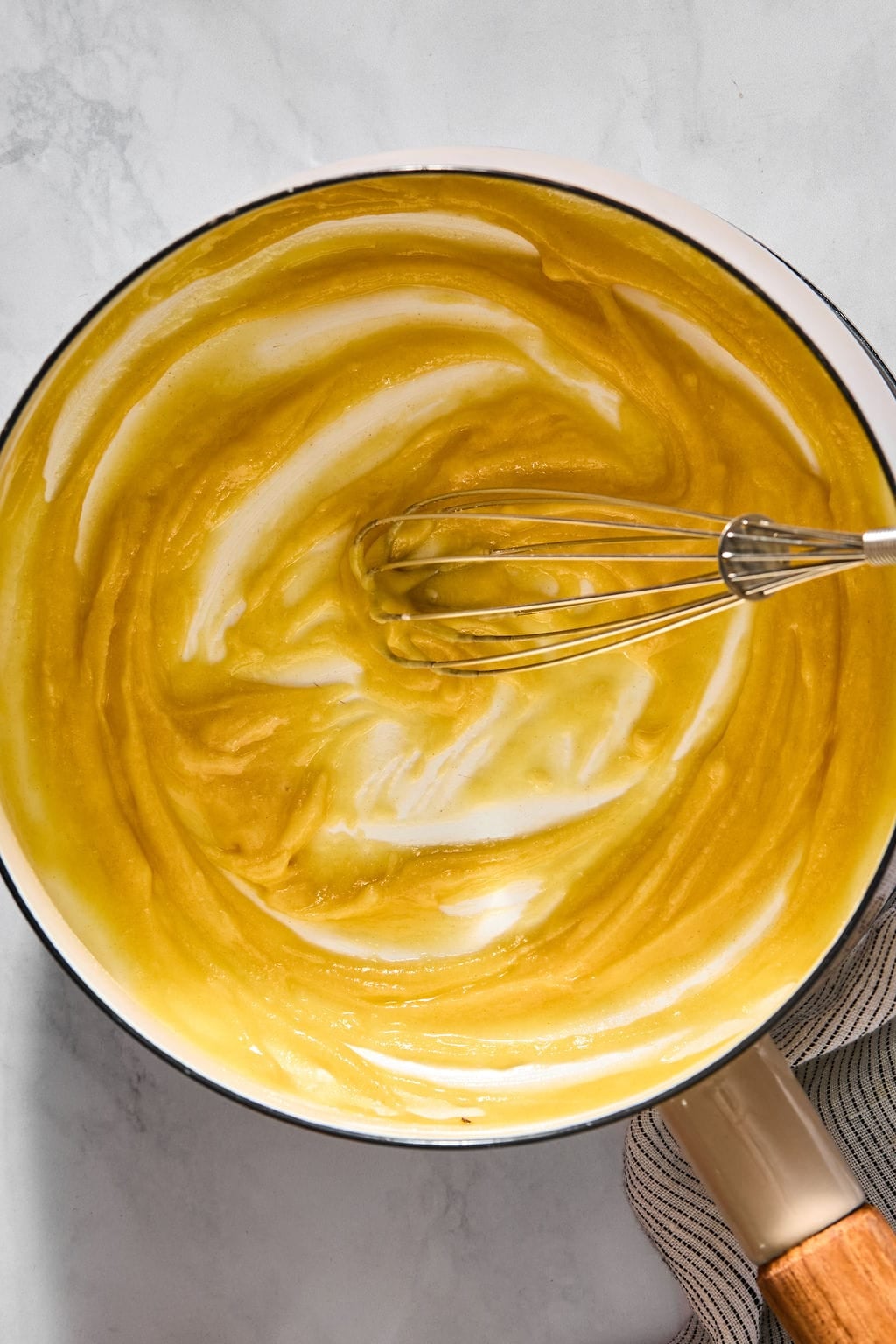A metal whisk stirring a smooth, golden-yellow sauce in a light-colored saucepan.