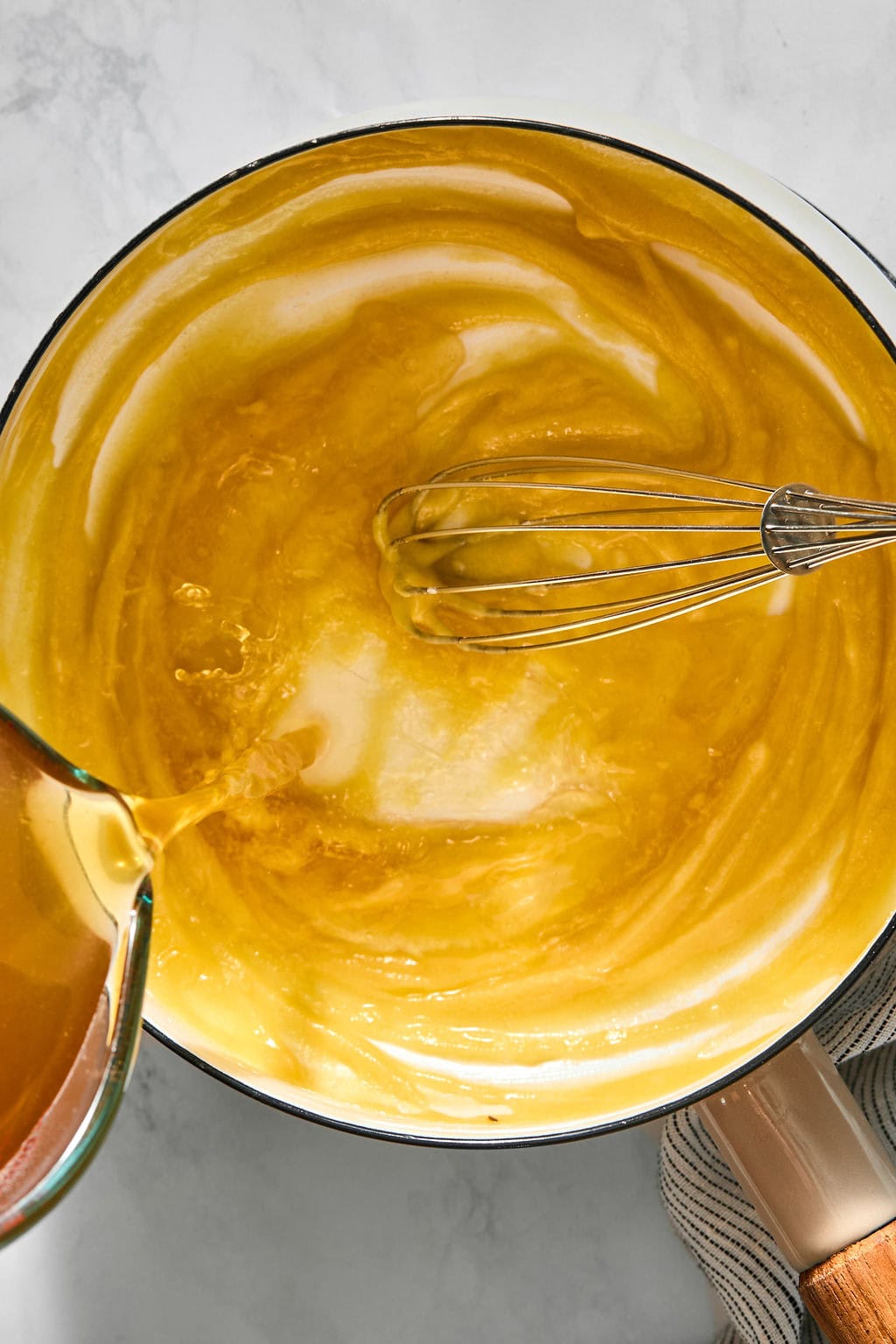 Liquid being poured from a glass measuring cup into a pot containing a yellow mixture, with a metal whisk inside.