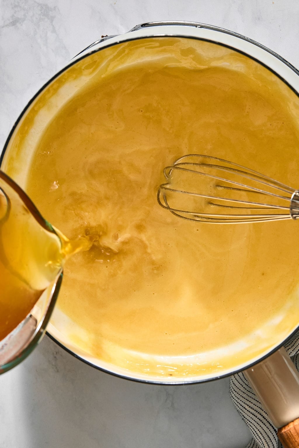 Liquid being poured from a measuring cup into a pot containing a yellow sauce and a metal whisk.