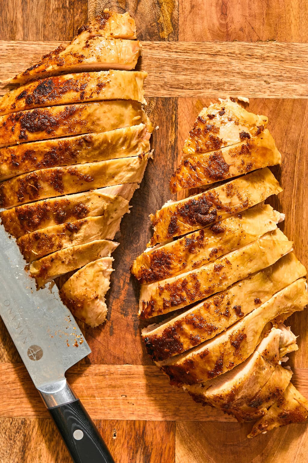 Two sliced, seasoned turkey breasts on a wooden cutting board with a chefs knife.