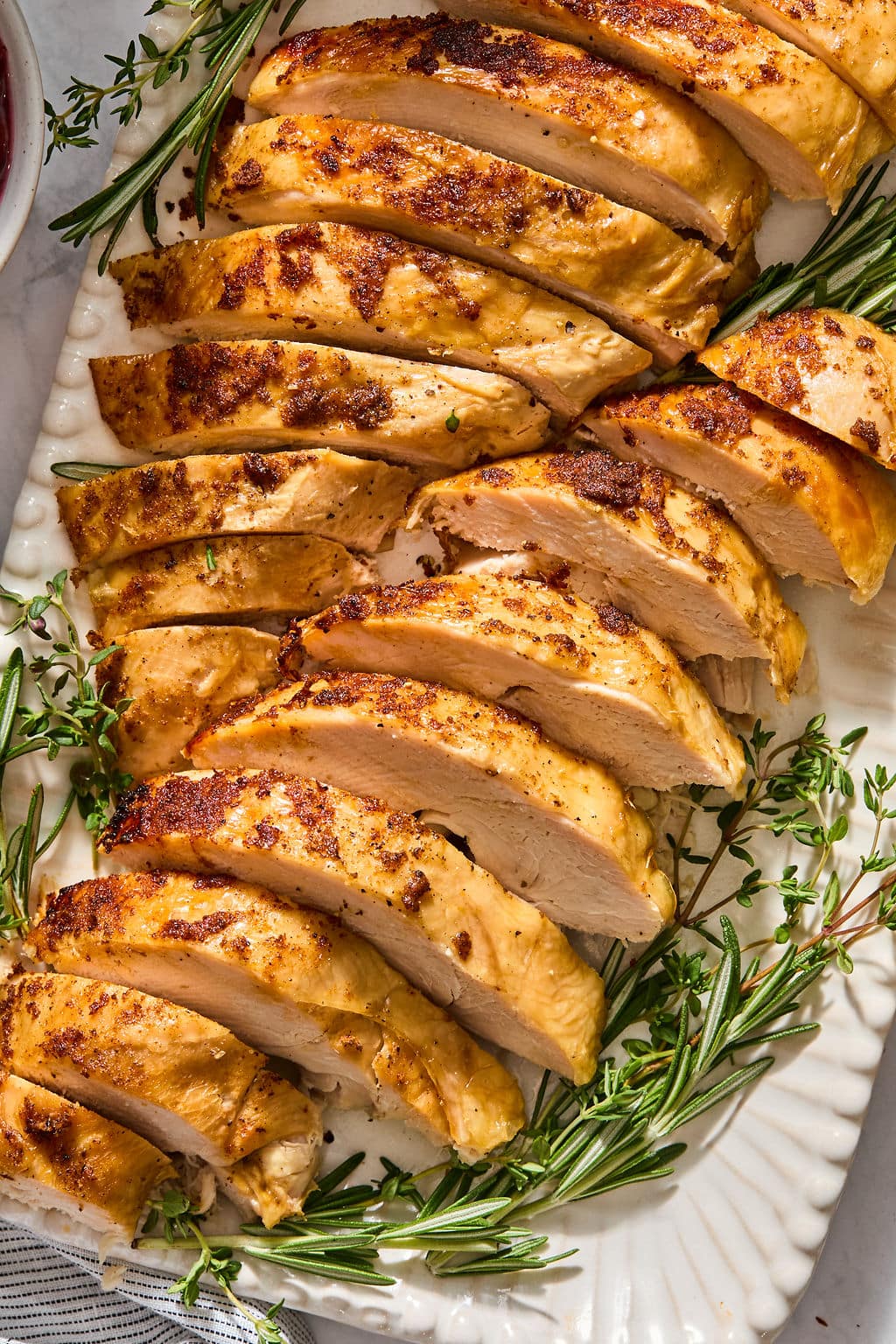 Sliced roasted turkey breast garnished with fresh rosemary and thyme on a white platter.