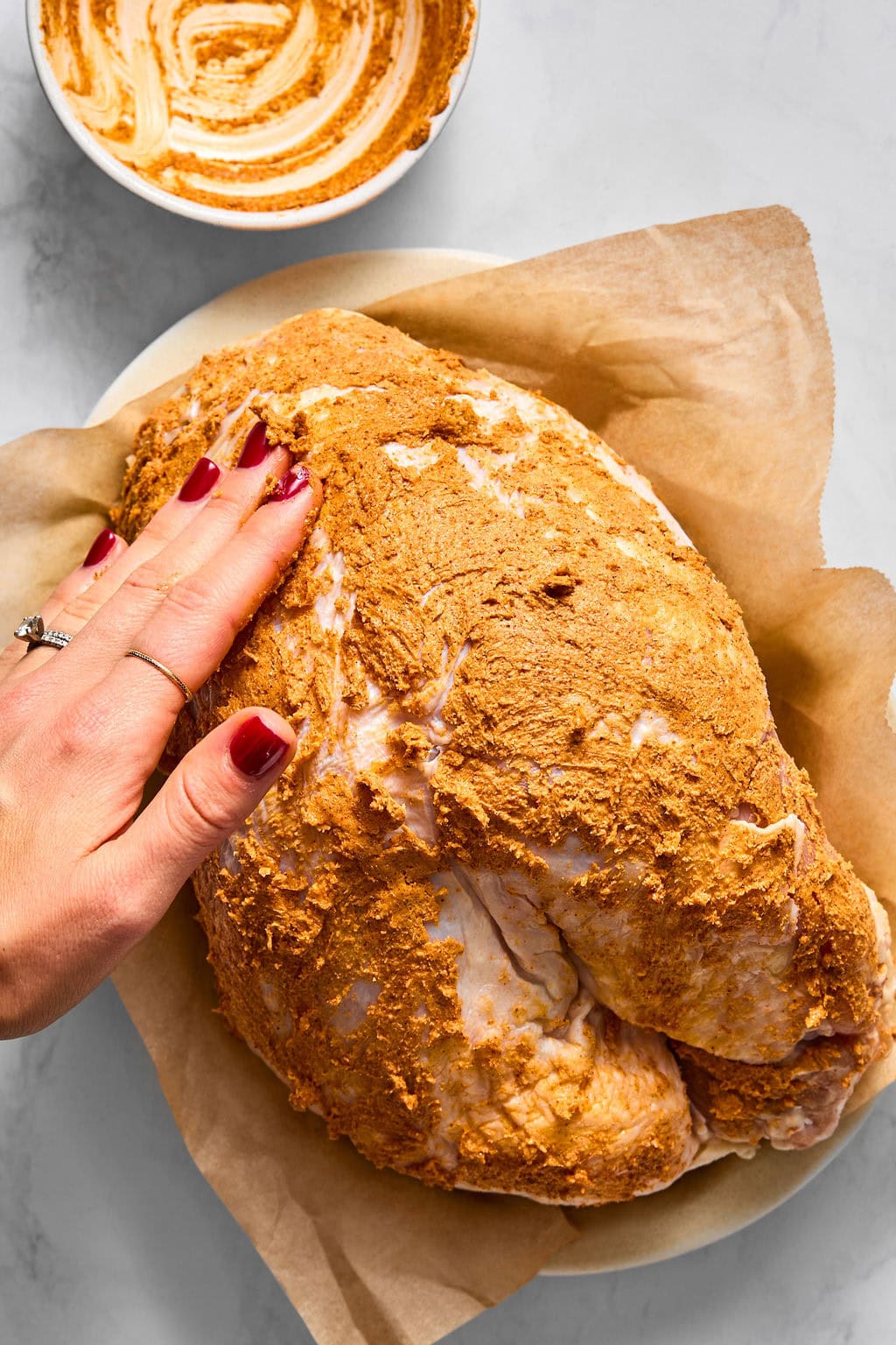 A hand rubs a dry spice rub onto a raw turkey breast on parchment paper, with a bowl of the spice mixture nearby.