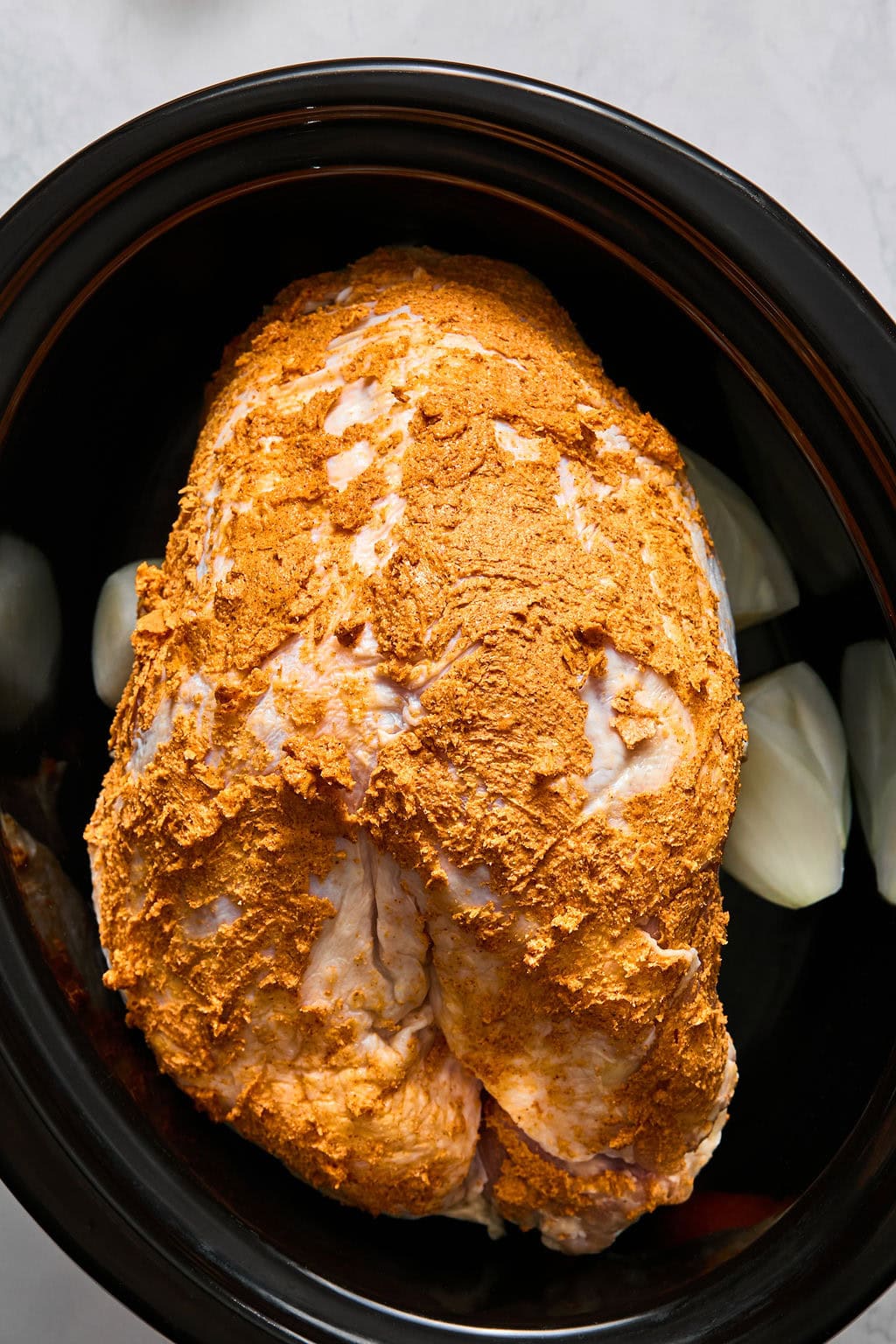 A raw turkey breast coated in dry orange seasoning inside a slow cooker with raw onion wedges.