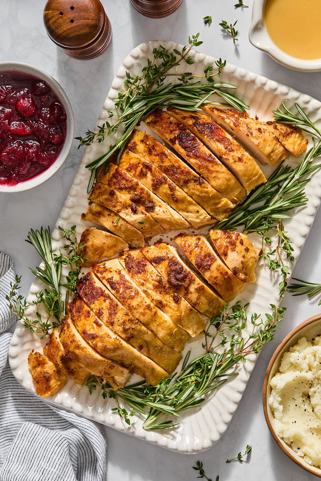 Sliced roasted turkey breast garnished with fresh rosemary and thyme on a rectangular platter, served with bowls of cranberry sauce and mashed potatoes.