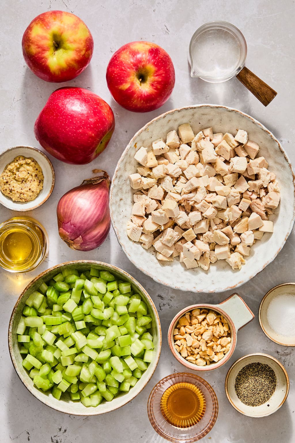 Ingredients for a salad arranged on a table: diced chicken, chopped celery, three red apples, a shallot, peanuts, mustard, oil, vinegar, and seasonings.