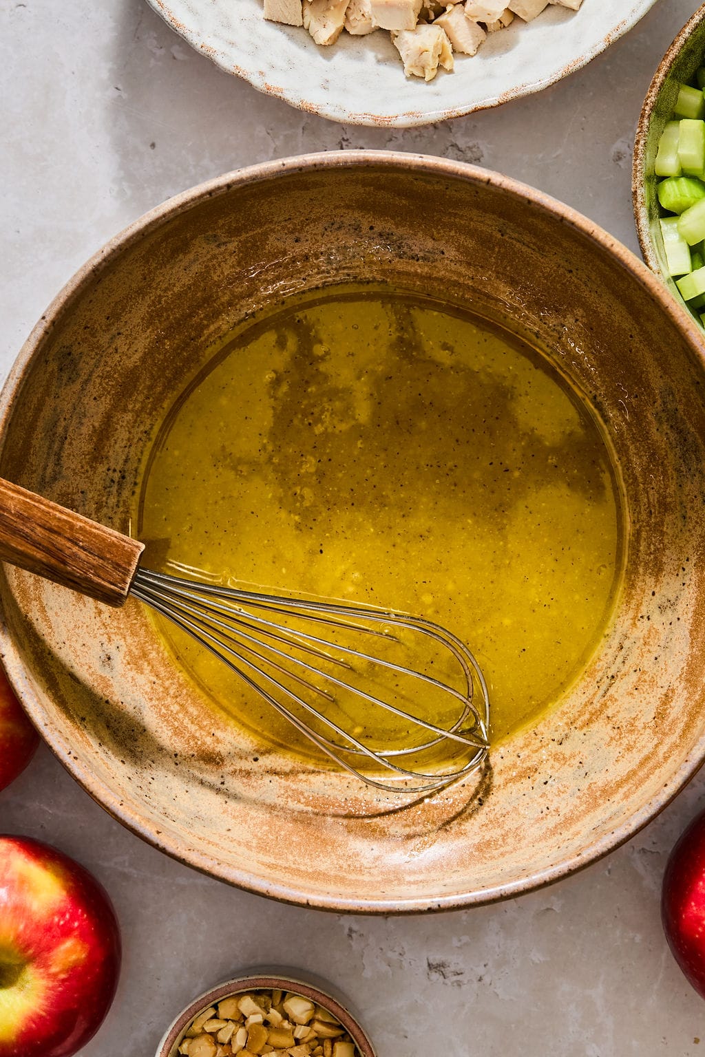 A metal whisk in a bowl of whisked yellow dressing, surrounded by ingredients including diced chicken, celery, apples, and nuts.