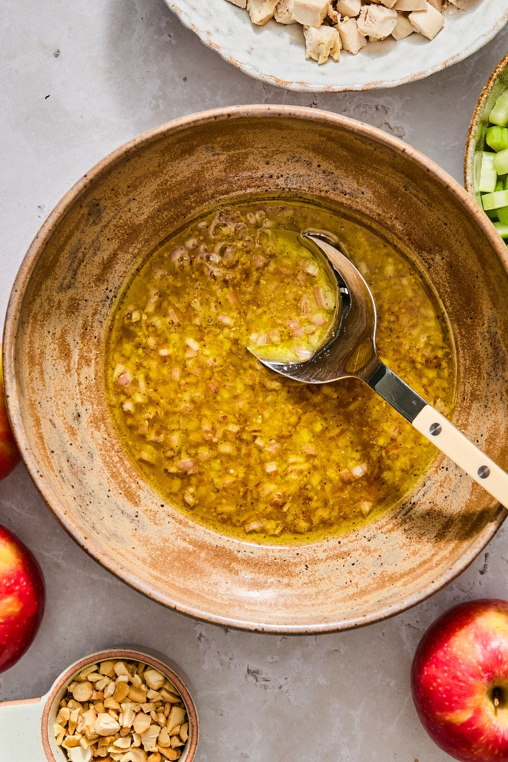 A metal spoon in a bowl of vinaigrette dressing, surrounded by small bowls of diced chicken, celery, and chopped nuts, with two red apples nearby.