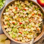 A bowl of chicken salad with diced apples, celery, and nuts, served with a metal spoon and whole apples nearby.