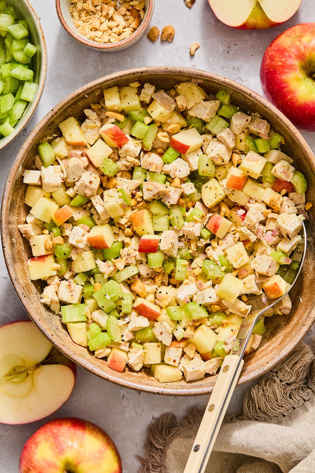 A bowl of chicken salad with diced apples, celery, and nuts, served with a metal spoon and whole apples nearby.