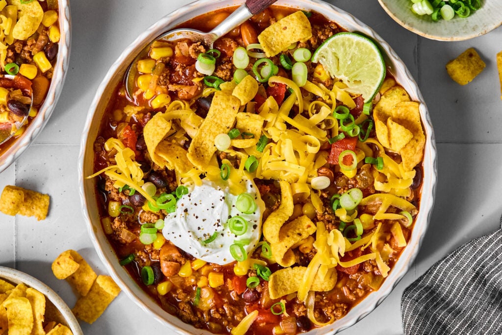 A bowl of taco soup topped with shredded cheese, a dollop of sour cream, corn chips, green onions, and a lime wedge.